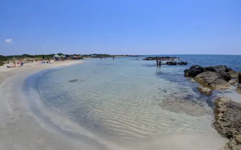 Spiaggia di sabbia bianca e mare cristallino a Marina di Salve, con scogli e persone che si godono il sole.
