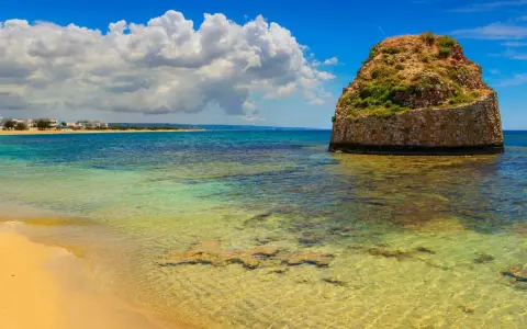 L'isola della fanciulla, una torre rotonda in pietra nel mare cristallino, con una spiaggia dorata e case in lontananza sotto un cielo azzurro con nuvole bianche.