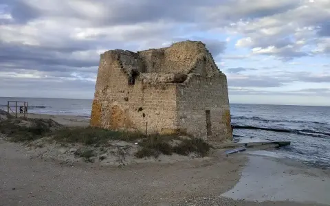 Ruined stone tower on a sandy beach with the sea and cloudy sky in the background.