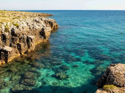 Torre solitaria su una scogliera rocciosa che domina un mare cristallino. La spiaggia sabbiosa è nascosta tra le rocce.