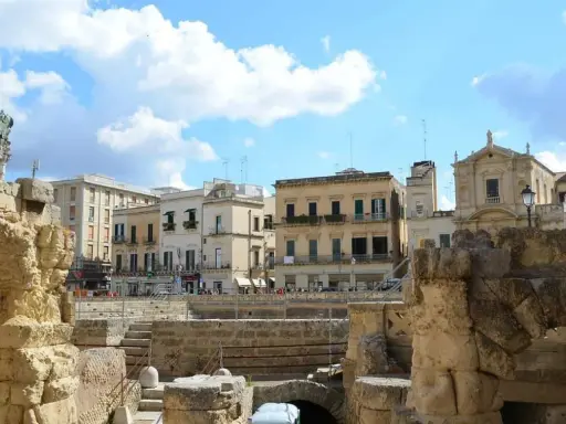 Antiche rovine romane si affacciano su palazzi barocchi nel centro storico di Lecce, sotto un cielo azzurro con nuvole bianche.