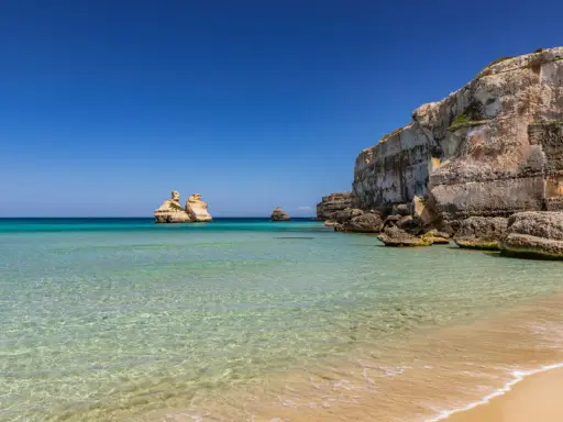 Spiaggia dorata e mare cristallino in Puglia, con faraglioni che emergono dalle acque turchesi e scogliere imponenti.