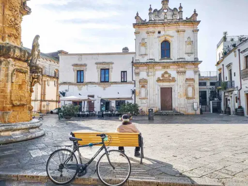 Un uomo seduto su una panchina nel **pescoluse centro storico**, con una bicicletta appoggiata. Sullo sfondo, una chiesa barocca e un caffè all'aperto.