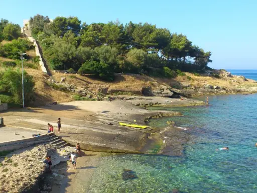 Scogliera e mare cristallino a San Gregorio, Puglia. Persone si godono il sole e l'acqua, con una scalinata che sale verso la vegetazione.