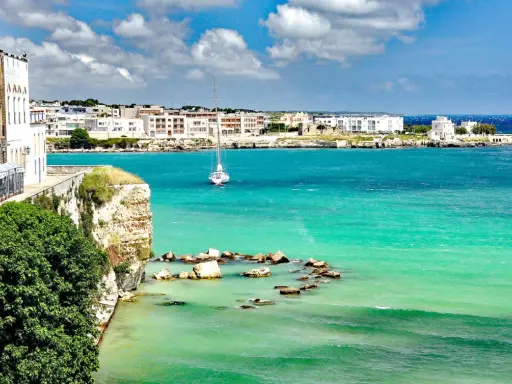 Vista mozzafiato del paese di Otranto con le sue case bianche affacciate sul mare turchese e un veliero solitario.