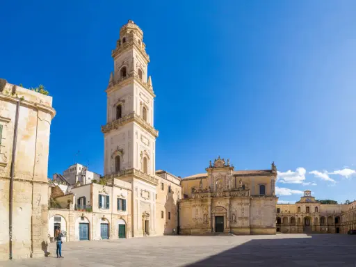La piazza Carmelo Bene a Lecce, con la maestosa Basilica di Santa Croce e il campanile che svetta sotto un cielo azzurro.