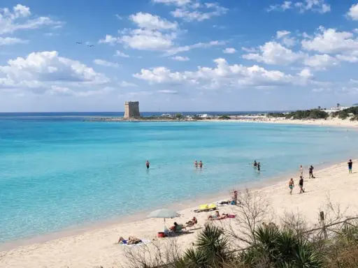 Spiaggia Scala di Furno con torre antica e acque turchesi. Persone si godono il sole e il mare cristallino.