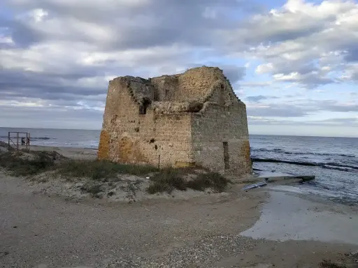 Ruined stone tower on a sandy beach with the sea and cloudy sky in the background.