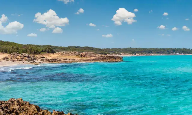 Acque cristalline e scogliere rocciose a Marina di Lecce, con una spiaggia sabbiosa e vegetazione mediterranea sullo sfondo sotto un cielo azzurro con nuvole bianche.