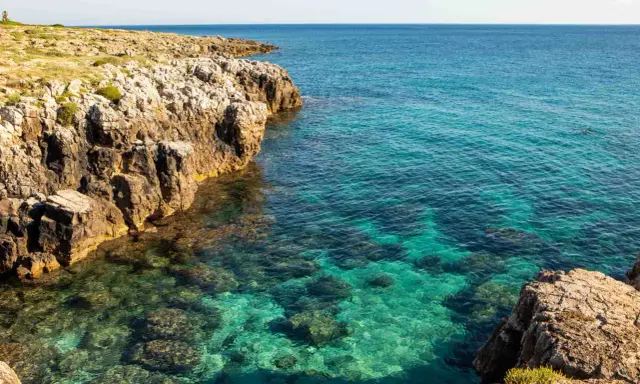 Torre solitaria su una scogliera rocciosa che domina un mare cristallino. La spiaggia sabbiosa è nascosta tra le rocce.