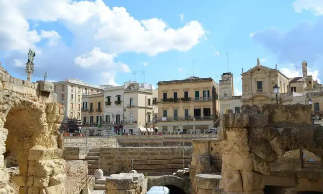 Antiche rovine romane si affacciano su palazzi barocchi nel centro storico di Lecce, sotto un cielo azzurro con nuvole bianche.