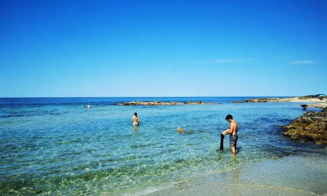 Spiaggia calette di Salve con gente che nuota e un cane che gioca nell'acqua cristallina sotto un cielo azzurro.