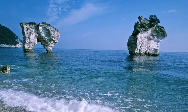 La **Baia delle Zagare** con le sue iconiche faraglioni bianchi che emergono dal mare blu intenso, sotto un cielo limpido.