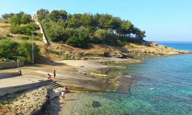 Scogliera e mare cristallino a San Gregorio, Puglia. Persone si godono il sole e l'acqua, con una scalinata che sale verso la vegetazione.