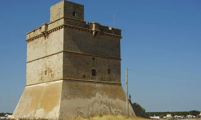 Torre costiera a Sant Isidoro, Salento. Antica sentinella di pietra contro il cielo azzurro, con il mare e il borgo sullo sfondo.