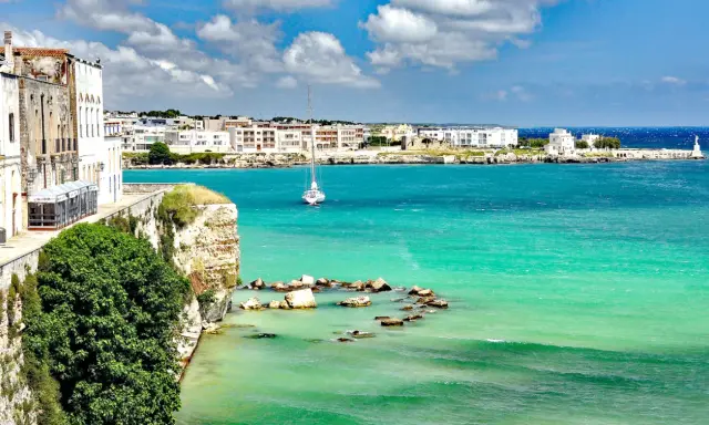 Vista mozzafiato del paese di Otranto con le sue case bianche affacciate sul mare turchese e un veliero solitario.