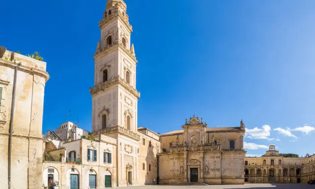 La piazza Carmelo Bene a Lecce, con la maestosa Basilica di Santa Croce e il campanile che svetta sotto un cielo azzurro.