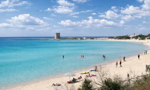 Spiaggia Scala di Furno con torre antica e acque turchesi. Persone si godono il sole e il mare cristallino.