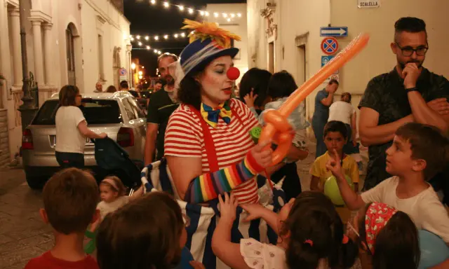 Clown con palloncino arancione anima la sagra di paese, circondata da bambini curiosi e adulti festanti.