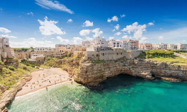 Un borgo bianco su scogliere a picco sul mare turchese, una spiaggia affollata e un invito a scoprire la Puglia da visitare.