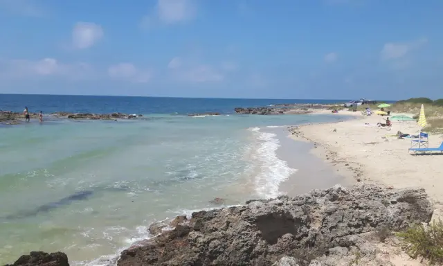 Spiaggia con scogli e mare cristallino, tipica delle calette puglia. Persone in acqua e sulla sabbia, ombrelloni e lettini.