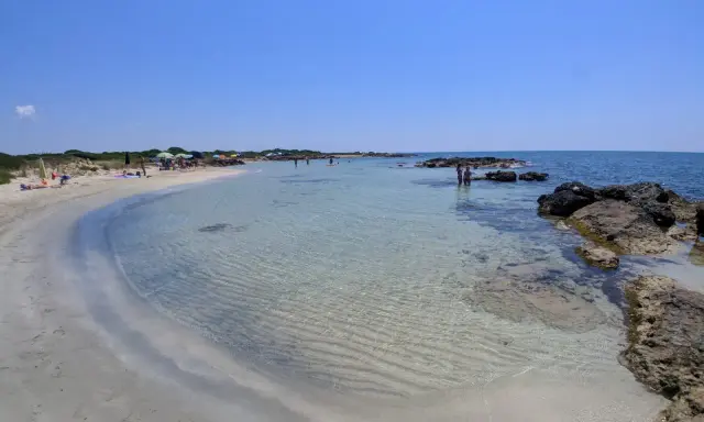 Spiaggia di sabbia bianca e mare cristallino a Marina di Salve, con scogli e persone che si godono il sole.