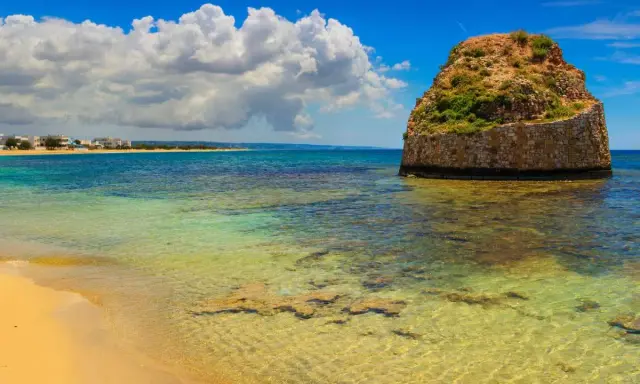 L'isola della fanciulla, una torre rotonda in pietra nel mare cristallino, con una spiaggia dorata e case in lontananza sotto un cielo azzurro con nuvole bianche.