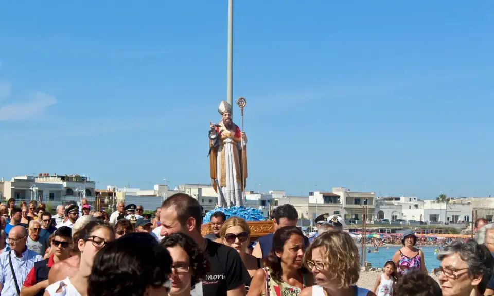 Festa San Foca: folla partecipa alla processione con la statua del santo su un carro, sotto un cielo azzurro.