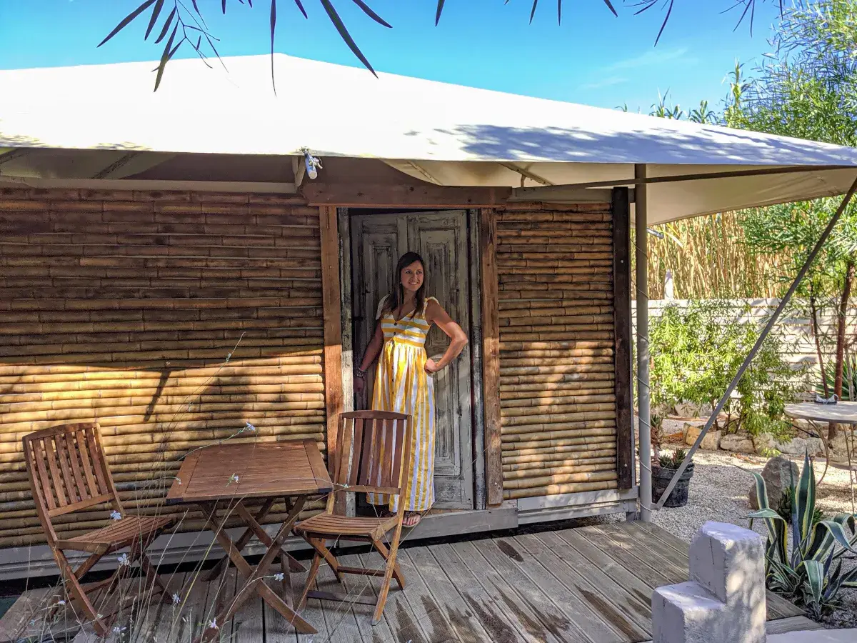 Donna sorridente in un abito a righe gialle davanti a una tenda di bambù, che incarna il glamping significato: lusso e natura.