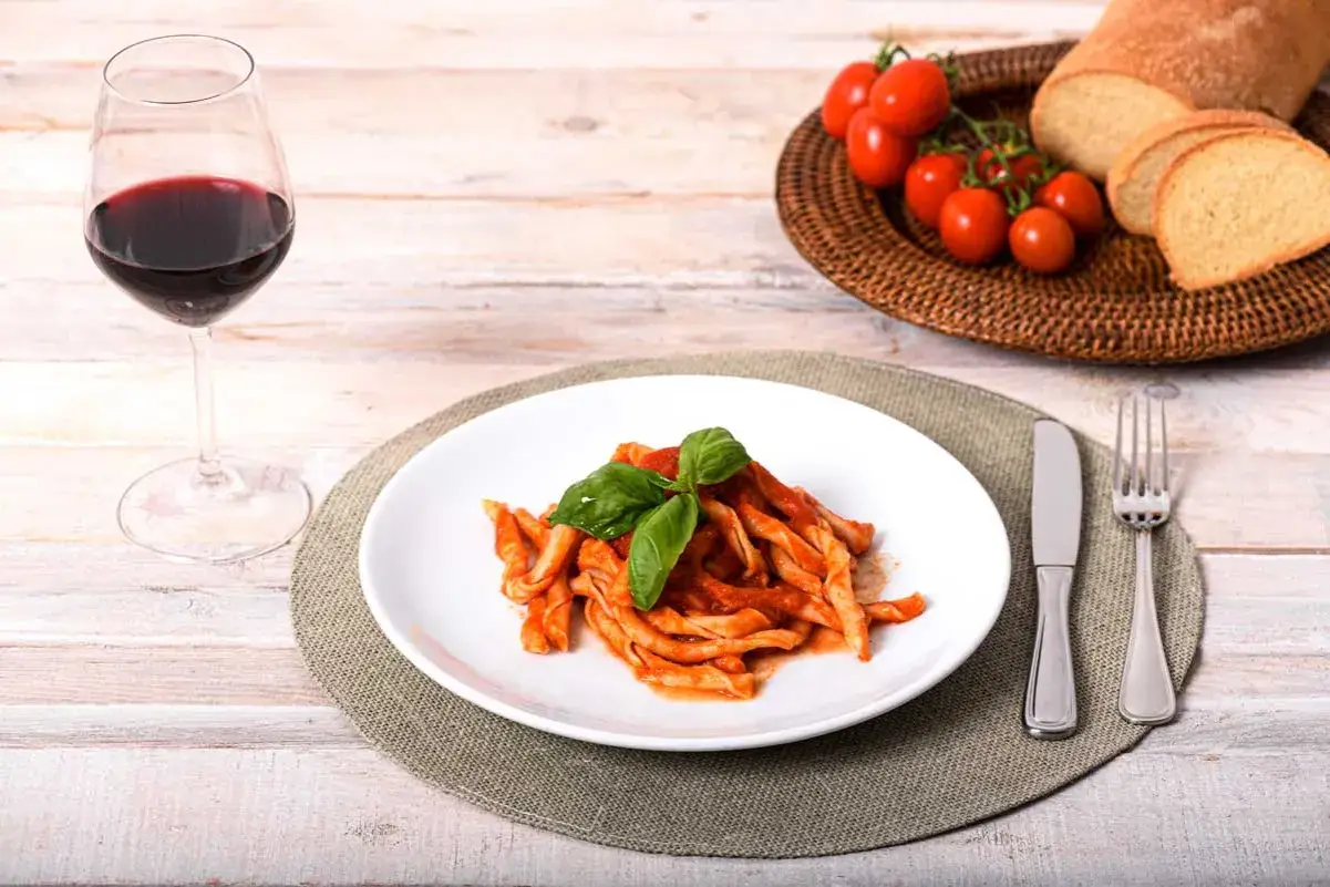 Piatto di sagne ncannulate al sugo, guarnite con basilico, accompagnate da un bicchiere di vino rosso, pane e pomodorini.