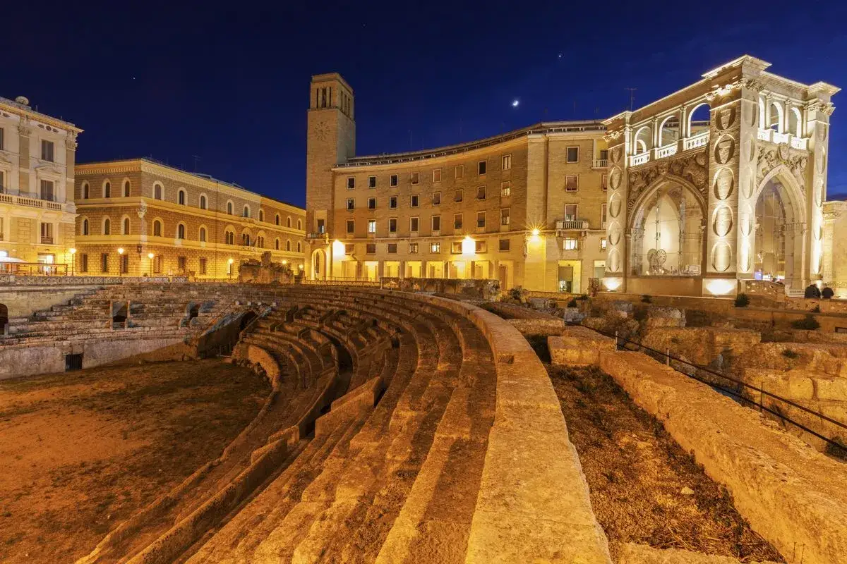 Antico anfiteatro romano illuminato di notte nel centro storico di Lecce, con edifici barocchi sullo sfondo.