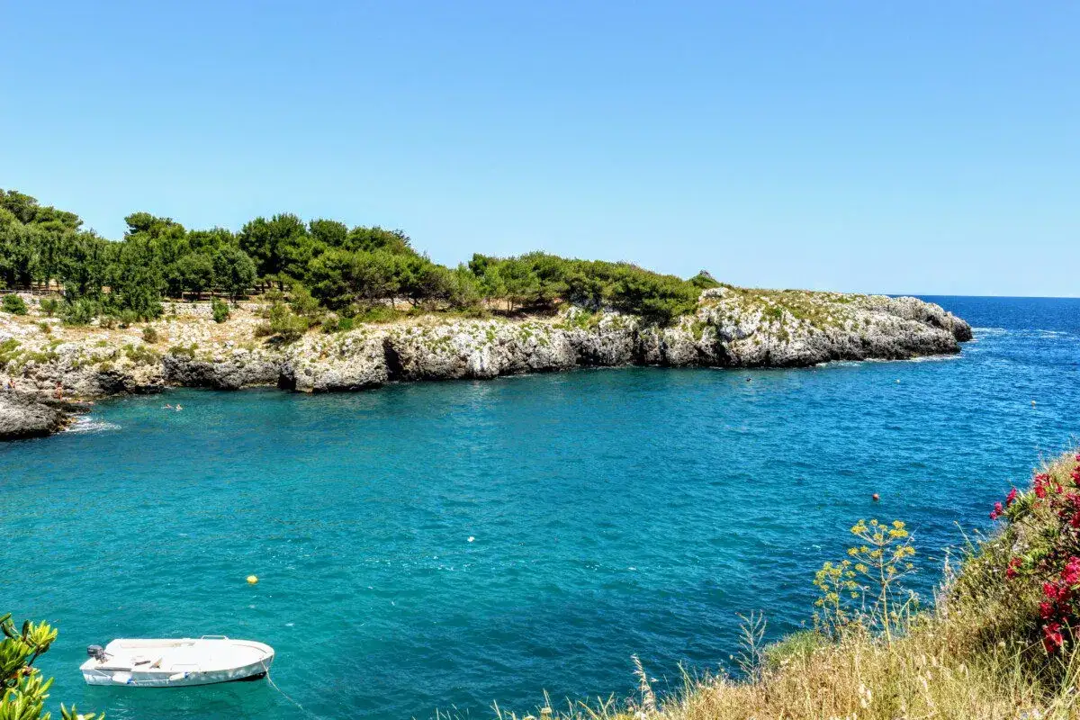 Acque cristalline e scogliere rocciose nel Salento Ionico. Una piccola barca bianca galleggia tranquilla, mentre il sole illumina la costa verdeggiante.