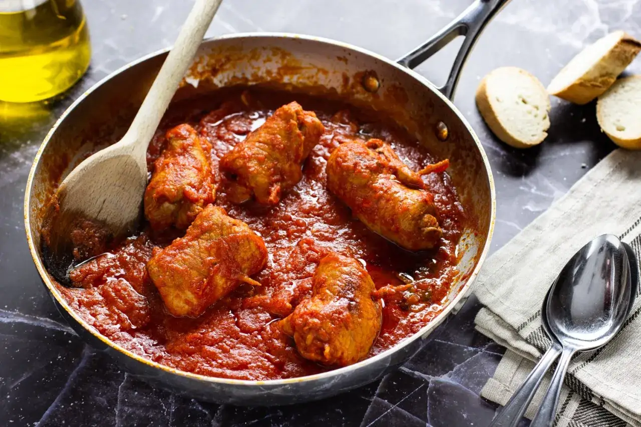 Braciola pugliese in padella con sugo rosso, accompagnata da pane e cucchiai.
