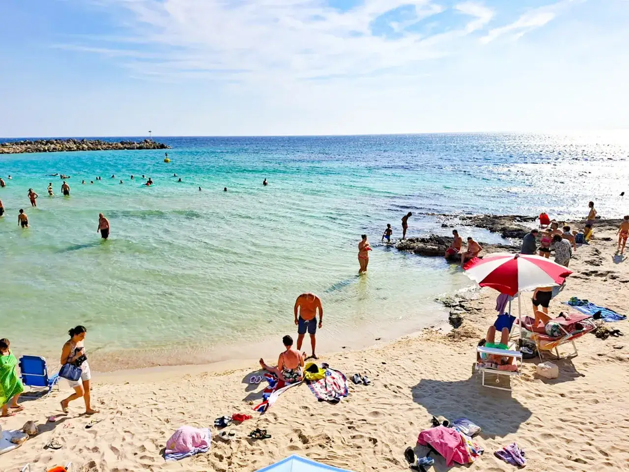 Spiaggia affollata con persone che nuotano e prendono il sole. In lontananza, una diga di rocce e il mare azzurro. La torre vado centro storico &egrave; un luogo ideale per godersi l'estate.