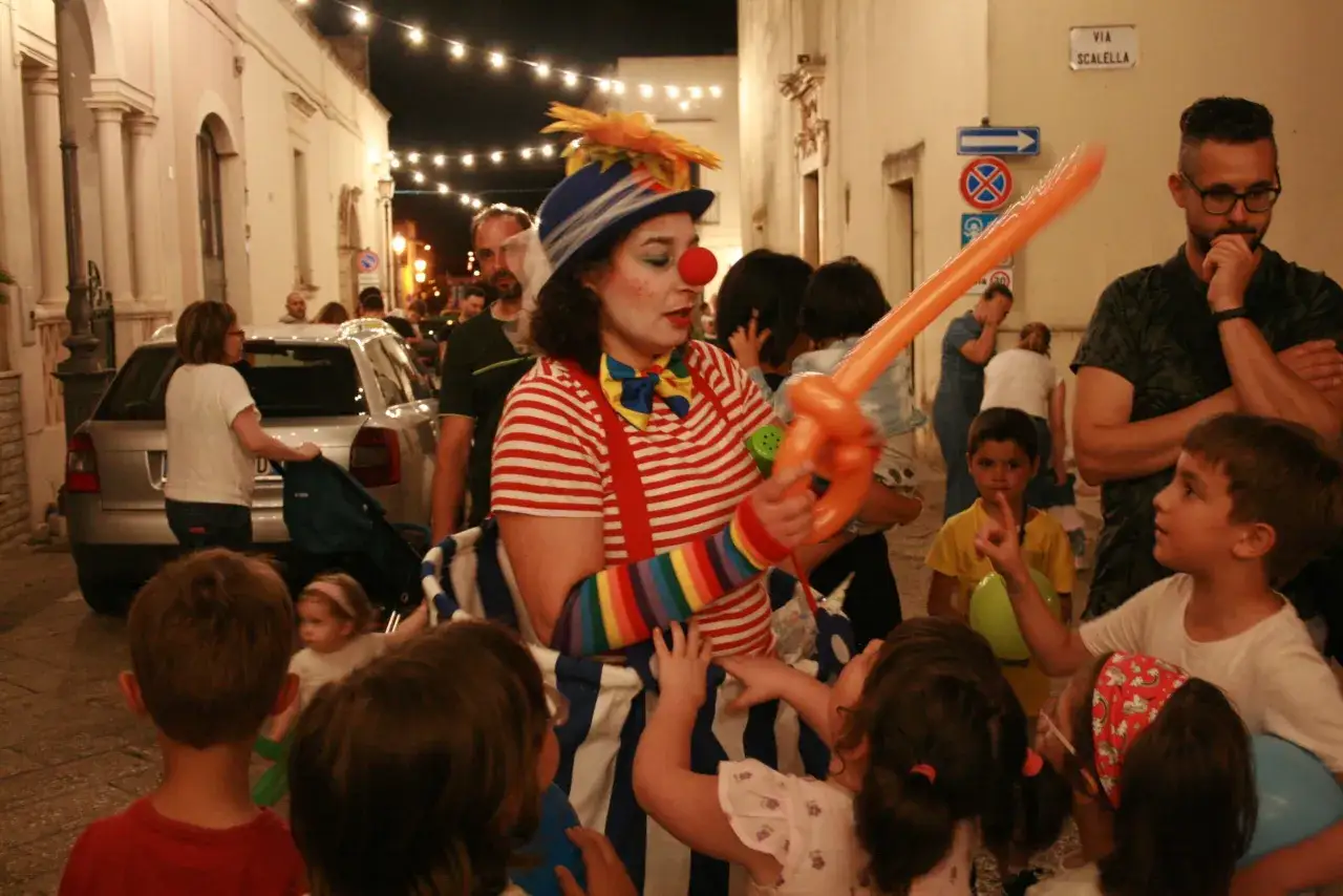 Clown con palloncino arancione anima la sagra di paese, circondata da bambini curiosi e adulti festanti.