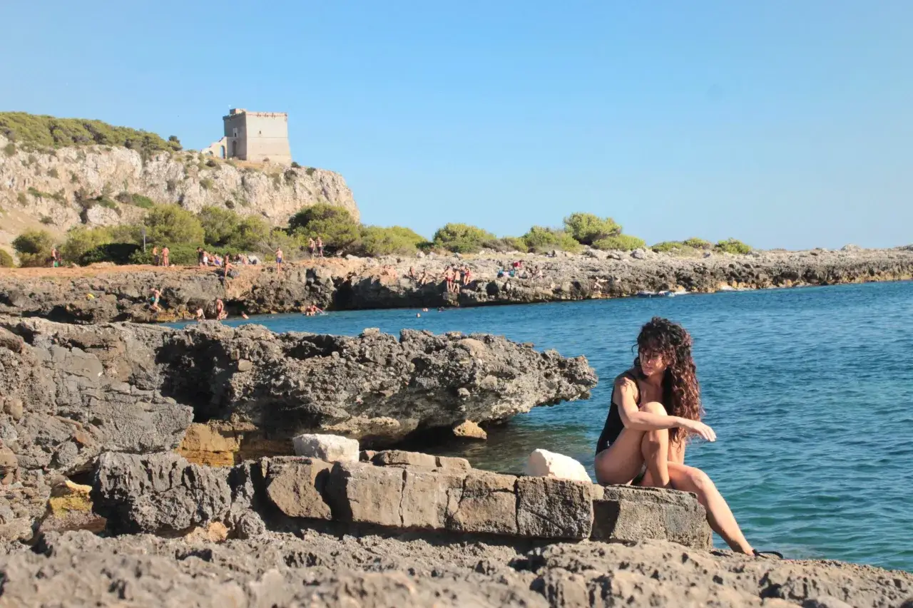 Donna con capelli ricci seduta su rocce, mare azzurro, torre in lontananza. Un'idea su cosa fare a Ferragosto in Puglia.