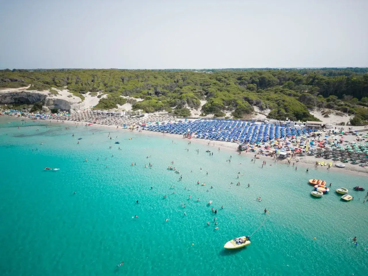 Vasta distesa di spiagge bianche affollate, con ombrelloni blu e verdi, bagnanti nell'acqua cristallina e una barca gialla.