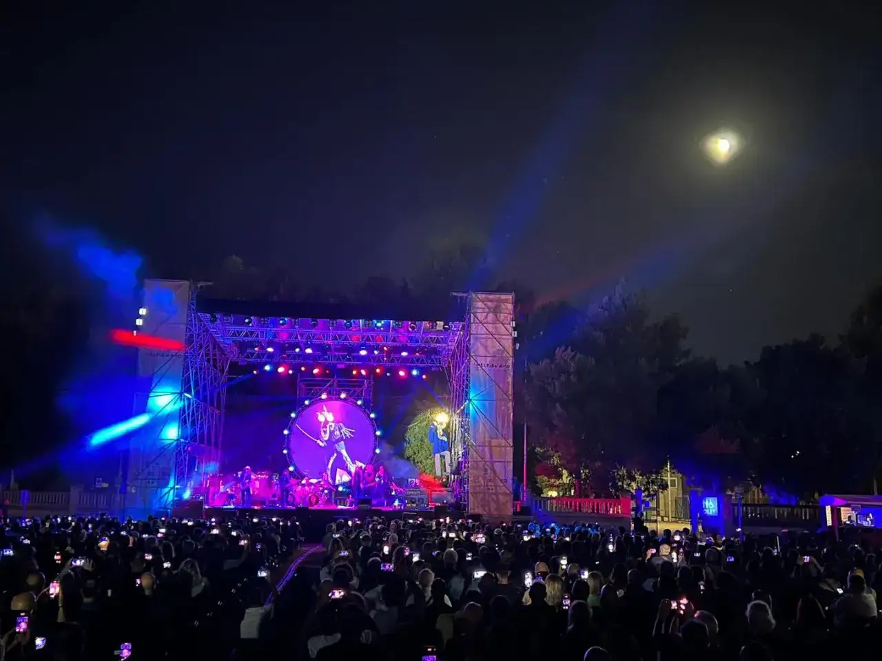 Un palco illuminato da luci blu e rosse, un grande schermo con un'immagine stilizzata, una folla di persone con i cellulari accesi. Magia dei concerti Salento sotto la luna piena.
