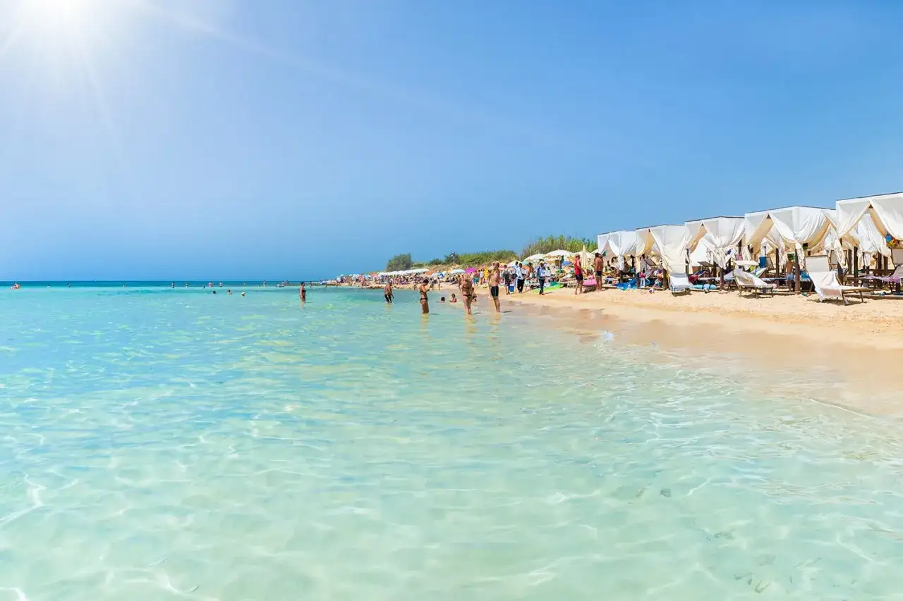 Spiagge sabbiose Puglia con acque cristalline e cabine bianche. Persone che si godono il sole e il mare in una giornata luminosa.