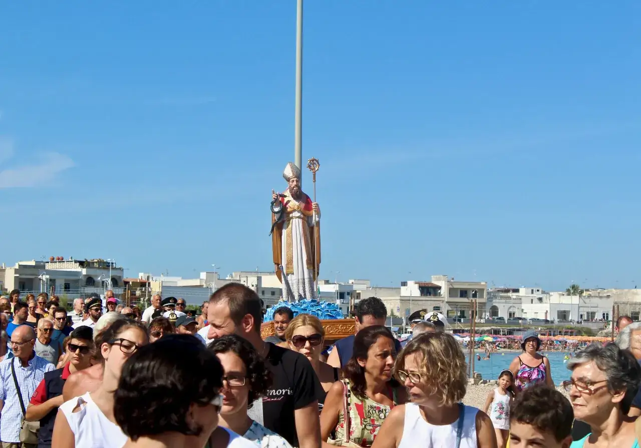 Festa San Foca: folla partecipa alla processione con la statua del santo, sullo sfondo il mare e le case.