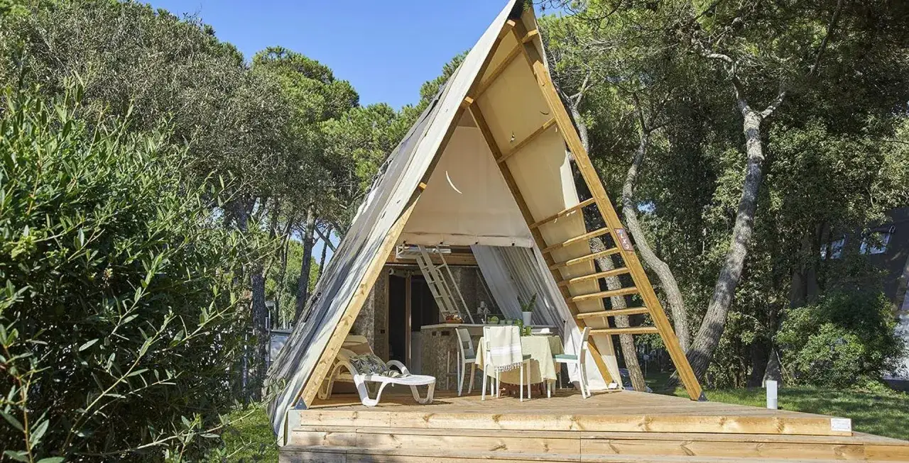 Struttura a forma di A in un bosco, con tavolo e sedie all'esterno. Questo è il glamping significato: lusso nella natura.