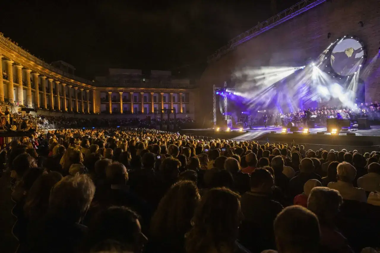 Un'orchestra e un coro si esibiscono sotto un cielo stellato, illuminati da fasci di luce viola. Il pubblico, numeroso, assiste con entusiasmo a uno dei tanti concerti Salento.