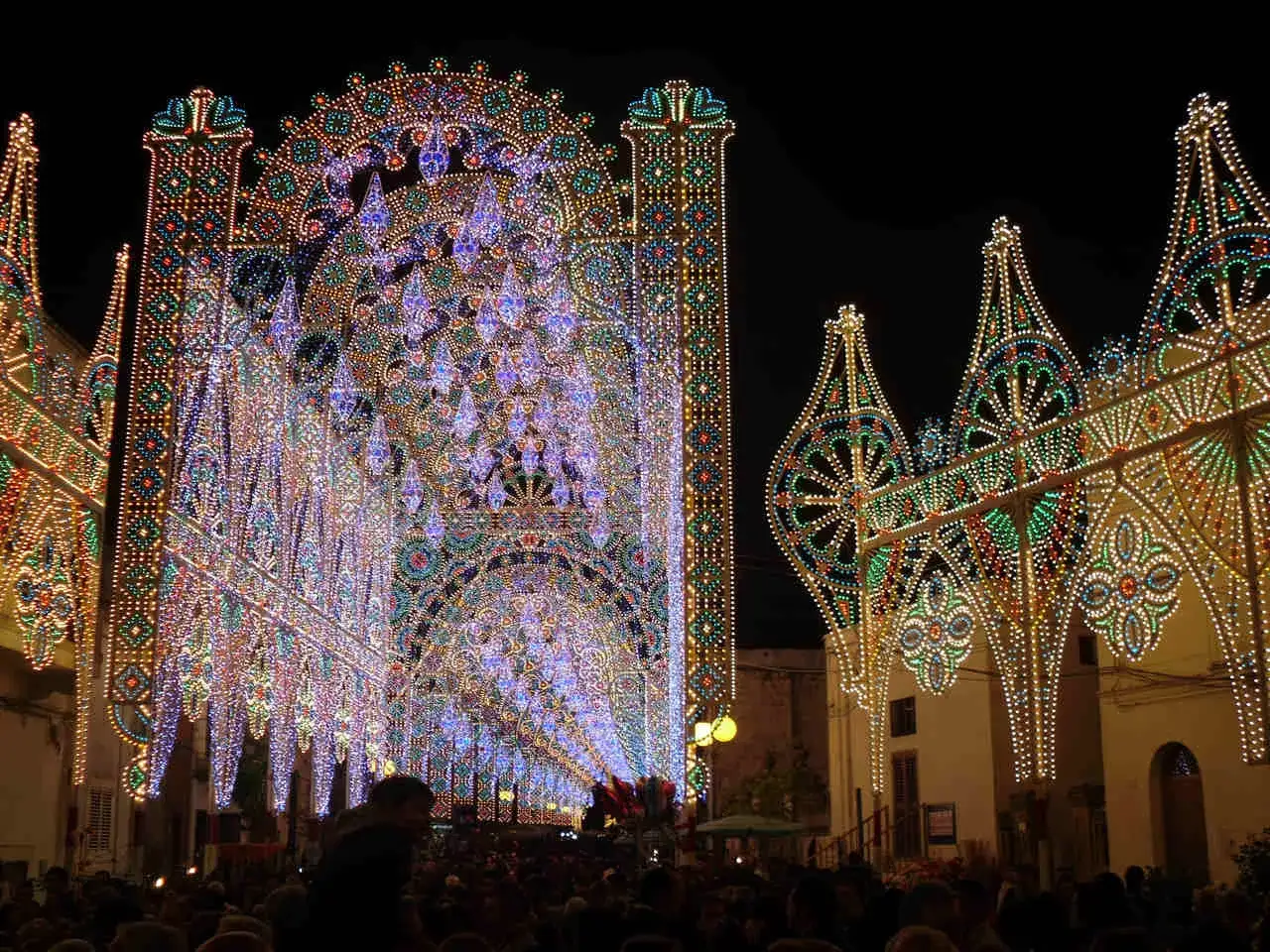 Un paese illuminato a festa per le sagre di paese. Archi di luci colorate creano un tunnel magico su una folla festante.
