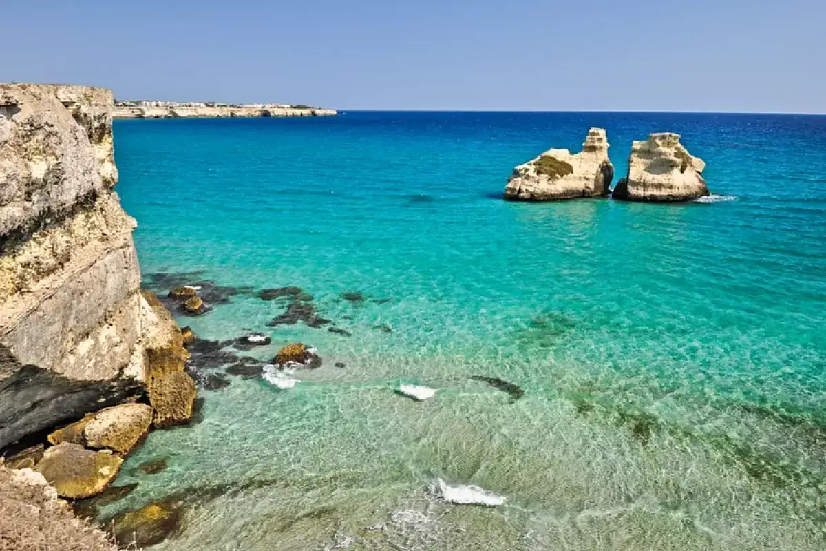Scogliera a picco sul mare cristallino, con faraglioni che emergono dall'acqua turchese. Una piccola spiaggia con sabbia dorata si intravede tra le rocce.