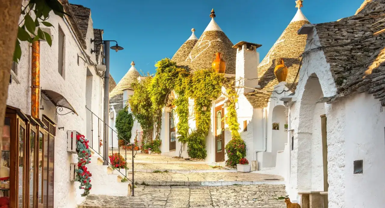 Un vicolo fiorito tra i trulli, tra i borghi pi&ugrave; belli d'Italia in Puglia. Un gatto arancione passeggia su un lastricato soleggiato.