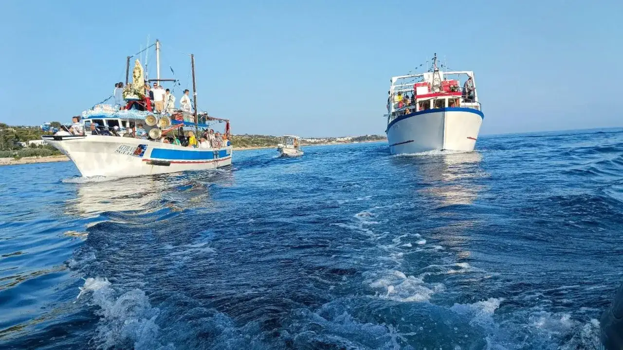 Barche in processione in mare per il Ferragosto Salento, con gente a bordo e una statua della Madonna.