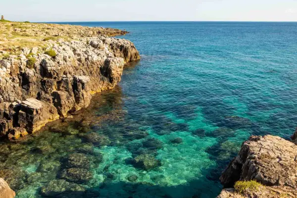 Torre solitaria su una scogliera rocciosa che domina un mare cristallino. La spiaggia sabbiosa è nascosta tra le rocce.