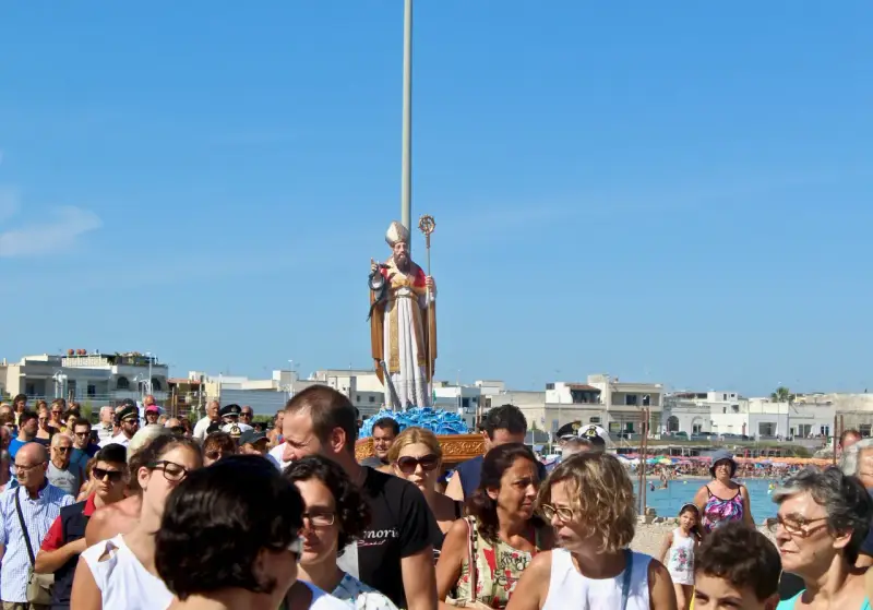 Festa San Foca: folla partecipa alla processione con la statua del santo su un carro, sotto un cielo azzurro.